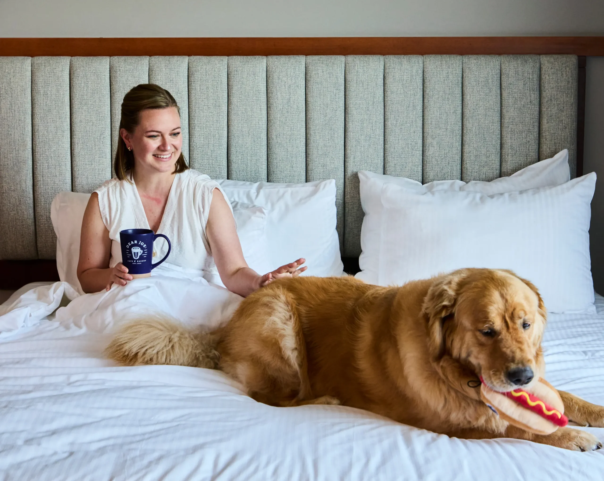 Woman with dog on bed, holding mug.